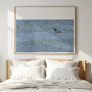 May include: A framed wall art print featuring a stylized illustration of a black and white loon swimming in blue water with white dashed ripples and patches of green reeds. The artwork is set against a plain white background.