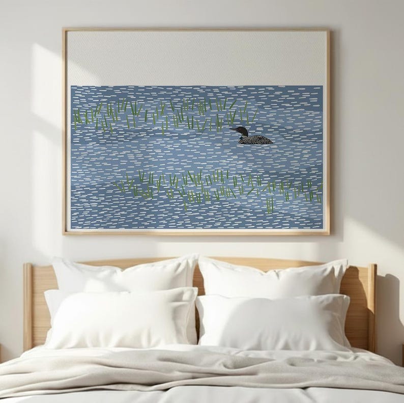 May include: A framed wall art print featuring a stylized illustration of a black and white loon swimming in blue water with white dashed ripples and patches of green reeds. The artwork is set against a plain white background.