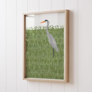 May include: A framed illustration of a grey heron standing in a field of green grass. The heron has an orange beak and black eyes.