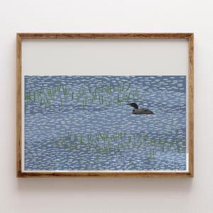 May include: A framed art print featuring a stylized illustration of a black and white loon swimming in blue water with small white dashes representing ripples and green vertical lines depicting aquatic plants in the foreground.