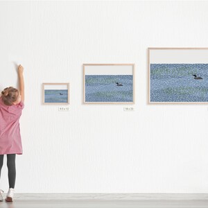 May include: Three framed wall art prints featuring a loon swimming on a blue patterned lake with green reeds. The frames are labeled with dimensions 9.5x12, 18x23, and 25x31 inches, displayed in a horizontal row on a white wall.