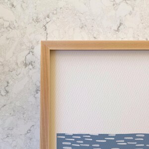 May include: A rectangular wall art piece featuring a light wood frame surrounding a textured off-white paper background. The lower portion of the artwork displays a horizontal band of blue with a repeating pattern of small white dashes.