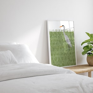 May include: A framed print of a grey heron standing in a field of green grass. The heron has an orange beak and is facing to the right. The print is in a white frame.