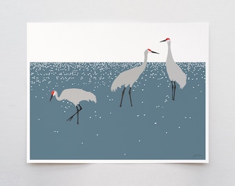 May include: A rectangular art print featuring three light gray sandhill cranes with red crowns and black legs standing in a dark blue body of water with white sparkling light reflections. The background is a solid white horizontal band.