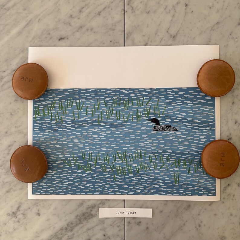 May include: A rectangular art print featuring a black and white loon swimming in blue water with white dashes and green reeds. The print is held by four round tan leather weights with the letters HJE embossed on them. The name JOREY HURLEY is printed below.