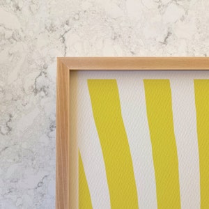 May include: A framed artwork with a pattern of vertical yellow and white stripes. The artwork is in a light-coloured wooden frame, adding a natural touch. The background is a textured, marble-like surface.