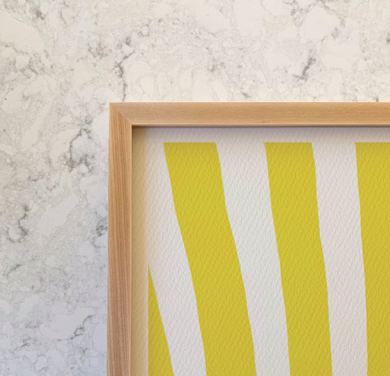 May include: A framed artwork with a pattern of vertical yellow and white stripes. The artwork is in a light-coloured wooden frame, adding a natural touch. The background is a textured, marble-like surface.