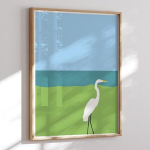 May include: A white egret stands on a green field with a blue body of water in the background. The sky is light blue.