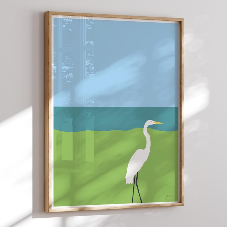 May include: A white egret stands on a green field with a blue body of water in the background. The sky is light blue.