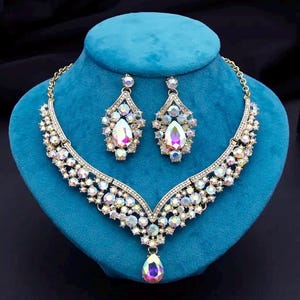 May include: A gold-toned necklace and matching drop earring set featuring iridescent, multi-faceted crystals. The V-shaped necklace has a central teardrop pendant, while the earrings mirror the design with a teardrop shape and crystal clusters.