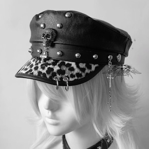 May include: Black leather cap with silver studs and a leopard print brim. The cap features a skull and cross pendant, along with silver hoop earrings. A gothic-inspired accessory.
