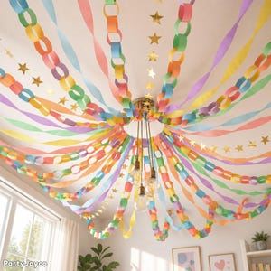 May include: A ceiling decoration featuring multiple paper chains in pastel rainbow colors, including pink, orange, yellow, green, blue, and purple. Gold star cutouts are interspersed among the chains, which radiate from a central light fixture.