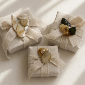 Puede incluir: Tres pequeñas cajas de regalo cuadradas envueltas en tela beige y atadas con una cinta a juego. Cada caja está adornada con un grupo de piedras naturales lisas de varios colores, incluyendo beige, marrón y negro, creando una estética rústica y elegante.