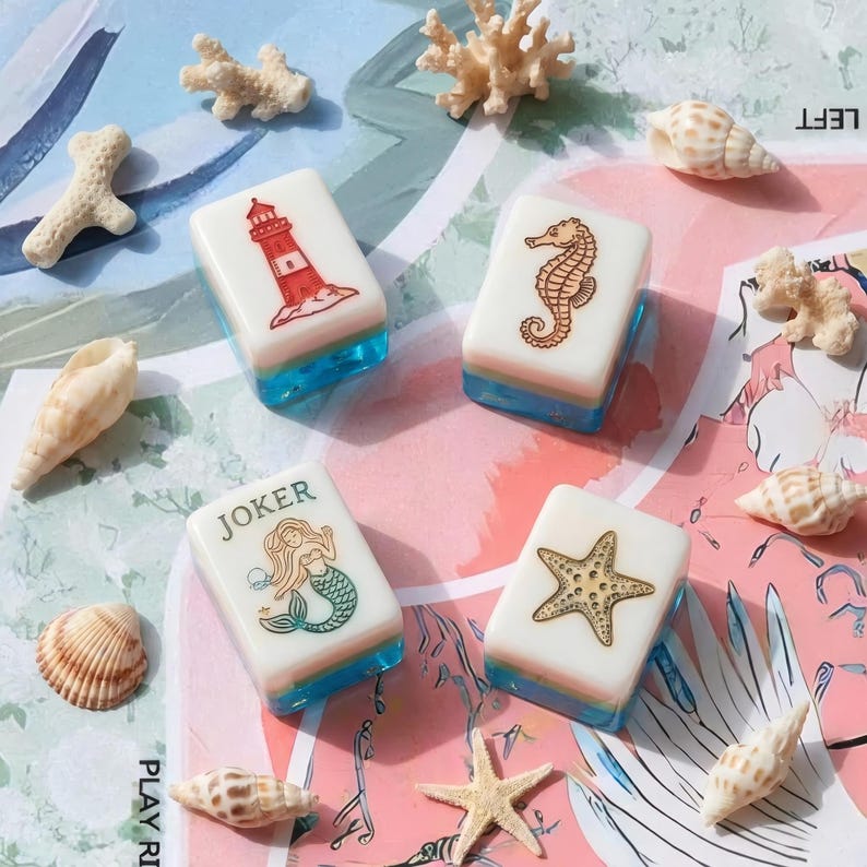 May include: Four rectangular mahjong tiles with white tops and translucent blue bases. The tiles feature engraved illustrations of a red lighthouse, a brown seahorse, a mermaid with the text JOKER, and a gold starfish, surrounded by various seashells.