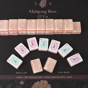 Puede incluir: Un juego de fichas de Mahjong con una fila de fichas de oro rosa en la parte superior y fichas blancas individuales con caracteres chinos coloridos y bordes florales debajo. El texto Mahjong Row & Co. est&aacute; impreso arriba con etiquetas.