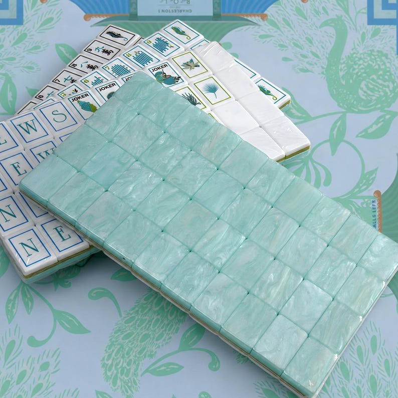 Puede incluir: Un conjunto de fichas de mahjong rectangulares que incluye una pila de fichas blancas con varios s&iacute;mbolos impresos en verde y negro, incluida la palabra JOKER, y una capa superior de fichas de color verde menta claro, lisas y veteadas, con acabado brillante.