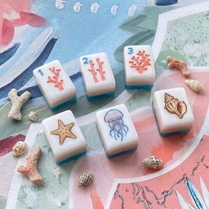May include: Six white rectangular mahjong tiles with blue bases, featuring illustrations of coral with numbers 1, 2, and 3, a starfish, a jellyfish, and a seashell, arranged alongside various small pieces of white and pink coral and seashells.