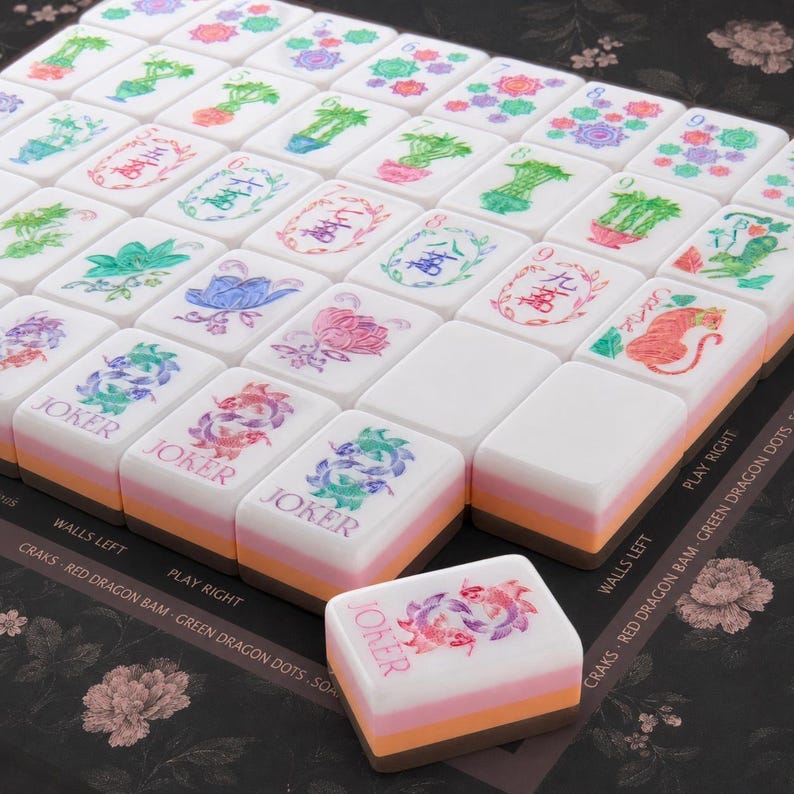 Puede incluir: Un juego de fichas de mahjong blancas con ilustraciones florales, num&eacute;ricas y de animales en varios colores. Cada ficha presenta un dise&ntilde;o en capas con una parte superior blanca, una central rosa y una base naranja. Varias fichas dicen JOKER.
