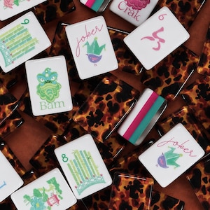 Könnte beinhalten: Eine Sammlung rechteckiger Mahjong-Spielsteine mit weißen Vorderseiten, die bunte Illustrationen von Bambus, Drachen und Joker-Figuren zeigen, sowie Spielsteine mit einem braun-schwarzen Schildpattmuster auf der Rückseite und den Kanten.