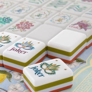 May include: A collection of rectangular Mahjong tiles featuring white tops with colorful illustrations of masks, flowers, and the word joker. Each tile has a layered side design with horizontal bands of white, olive green, and orange colors.