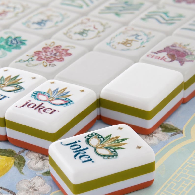 May include: A collection of rectangular Mahjong tiles featuring white tops with colorful illustrations of masks, flowers, and the word joker. Each tile has a layered side design with horizontal bands of white, olive green, and orange colors.