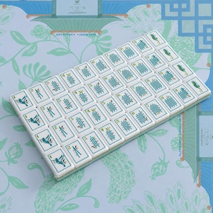Puede incluir: Un conjunto de fichas de mahjong rectangulares blancas dispuestas en cuadr&iacute;cula, con gr&aacute;ficos en color verde azulado y dorado de colibr&iacute;es, tallos de bamb&uacute; y caballitos de mar, cada una marcada con un peque&ntilde;o n&uacute;mero en la esquina.