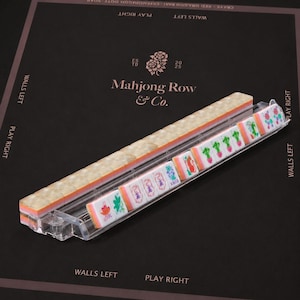 Puede incluir: Un soporte de pl&aacute;stico transparente que sostiene una fila de fichas de mahjong. Las fichas presentan una cara blanca con dise&ntilde;os florales y caracteres coloridos, incluida una ficha con la palabra Joker, y un reverso transl&uacute;cido marmoleado.