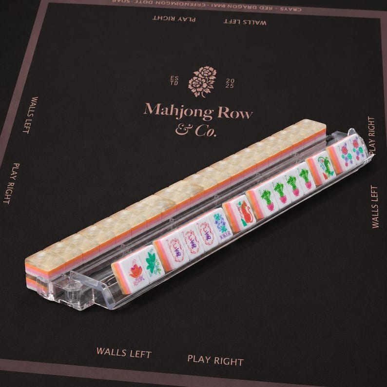 Puede incluir: Un soporte de pl&aacute;stico transparente que sostiene una fila de fichas de mahjong. Las fichas presentan una cara blanca con dise&ntilde;os florales y caracteres coloridos, incluida una ficha con la palabra Joker, y un reverso transl&uacute;cido marmoleado.