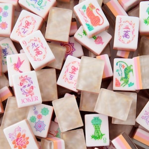 Puede incluir: Una colecci&oacute;n de fichas de mahjong rectangulares que presentan una mezcla de caras blancas con ilustraciones coloridas de animales, flores y caracteres chinos, junto a fichas beige jaspeadas transl&uacute;cidas con bordes en capas de color rosa y naranja.