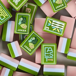 May include: A collection of rectangular mahjong tiles featuring a mix of pink, white, and green color schemes. Some tiles display the word JOKER with a graphic, others show the word EAST with an arrow, and some feature numerical symbols.