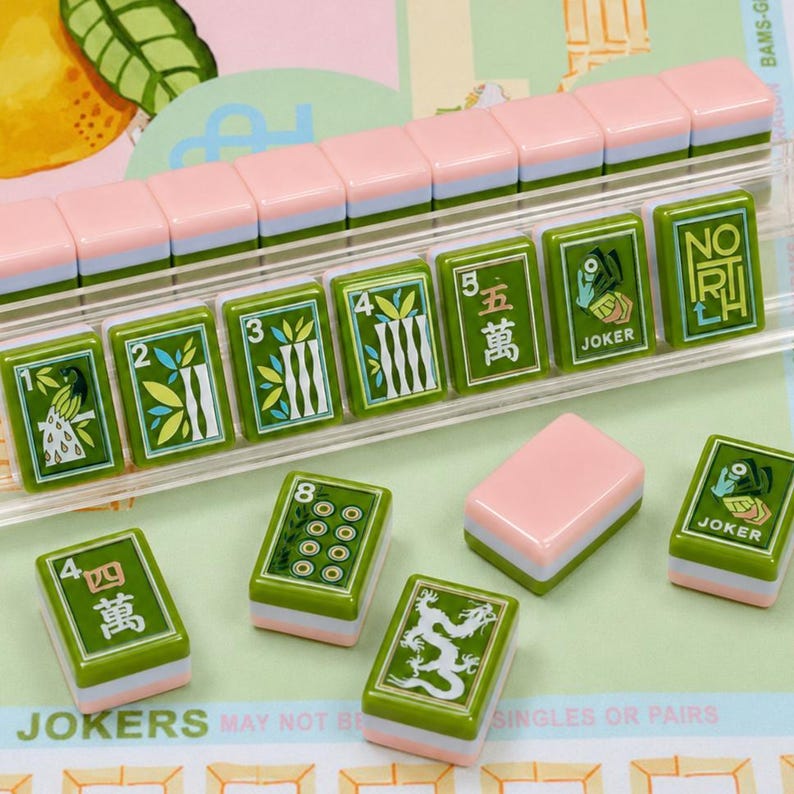 May include: A set of rectangular mahjong tiles featuring green faces with white and gold illustrations of numbers, bamboo, dragons, and the word JOKER, paired with pink and white bases. Some tiles are arranged in a clear plastic rack.