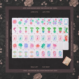 Puede incluir: Un juego de fichas de mahjong dispuestas en cuadr&iacute;cula sobre una alfombrilla floral oscura. Las fichas presentan dise&ntilde;os coloridos de flores, bamb&uacute;, n&uacute;meros y caracteres, incluyendo cinco fichas de Joker. El tapete tiene bordes con texto impreso.