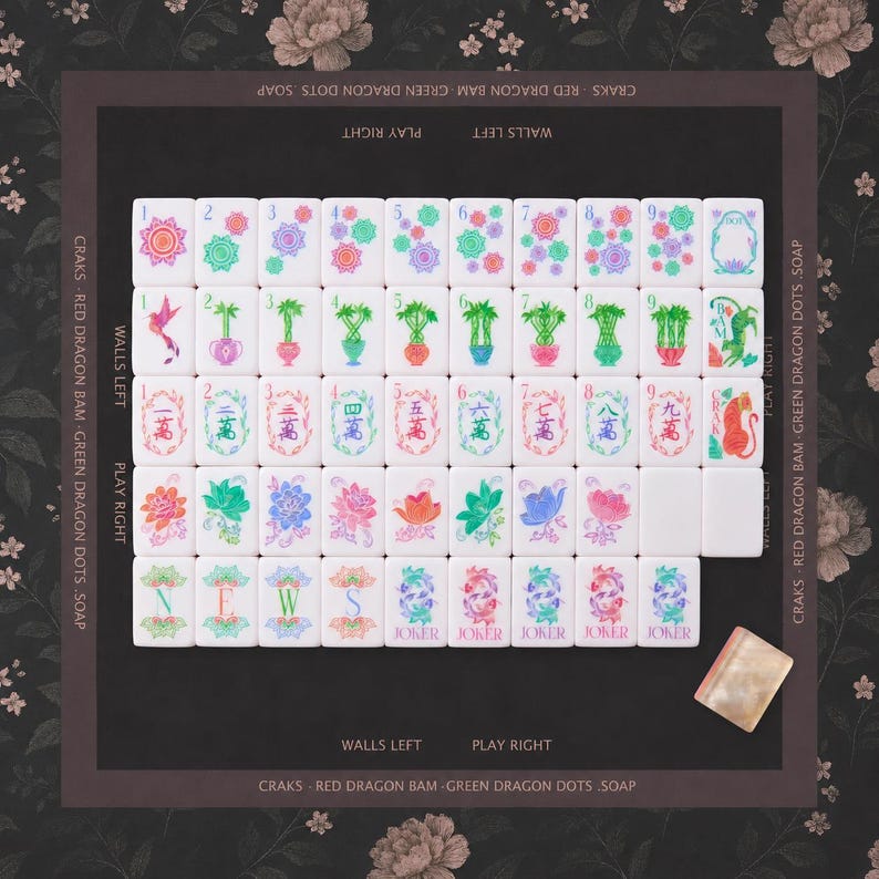 Puede incluir: Un juego de fichas de mahjong dispuestas en cuadr&iacute;cula sobre una alfombrilla floral oscura. Las fichas presentan dise&ntilde;os coloridos de flores, bamb&uacute;, n&uacute;meros y caracteres, incluyendo cinco fichas de Joker. El tapete tiene bordes con texto impreso.