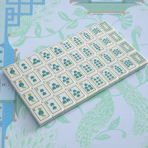 Puede incluir: Un conjunto rectangular de fichas de mahjong blancas dispuestas en una cuadr&iacute;cula, con dise&ntilde;os florales y num&eacute;ricos en azul. Cada ficha muestra un n&uacute;mero o s&iacute;mbolo espec&iacute;fico en tinta azul, con un acabado liso y brillante en la superficie.