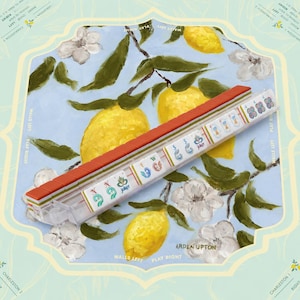 May include: A mahjong rack holding a row of white tiles with colorful symbols, including the word Joker, and red backs. The rack is positioned over a light blue background featuring a painted pattern of yellow lemons and white flowers.