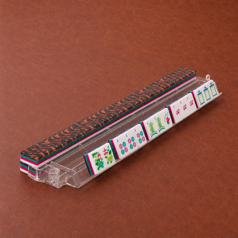 May include: A clear plastic mahjong tile rack holding a row of white tiles with green and pink engraved symbols, including bamboo and character designs, positioned next to a stack of tiles with a dark brown and black tortoiseshell pattern on the back.