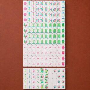 May include: A complete set of white Mahjong tiles arranged in a grid. The tiles feature colorful engraved designs including bamboo stalks, Chinese characters, floral patterns, directional letters N, E, W, S, and tiles labeled Joker, Bam, and Crak.
