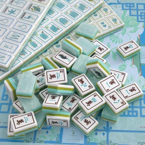 Puede incluir: Un juego de fichas de Mahjong con caras blancas y dise&ntilde;os en tinta azul y verde, que incluyen caracteres chinos, n&uacute;meros e ilustraciones de caballitos de mar y flamencos. Las fichas tienen un acabado veteado en verde claro y blanco.
