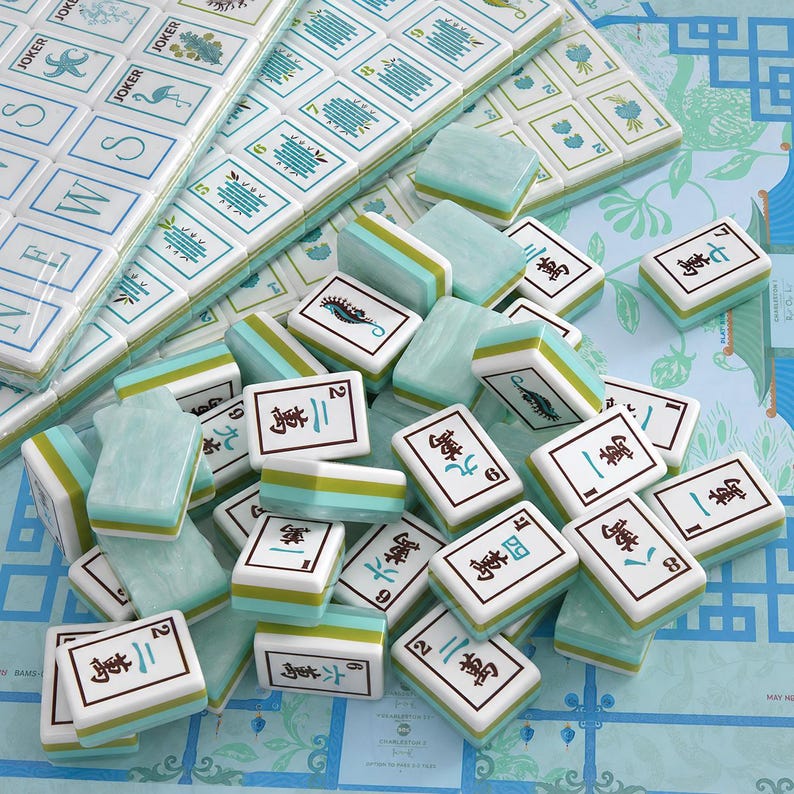 Puede incluir: Un juego de fichas de Mahjong con caras blancas y dise&ntilde;os en tinta azul y verde, que incluyen caracteres chinos, n&uacute;meros e ilustraciones de caballitos de mar y flamencos. Las fichas tienen un acabado veteado en verde claro y blanco.