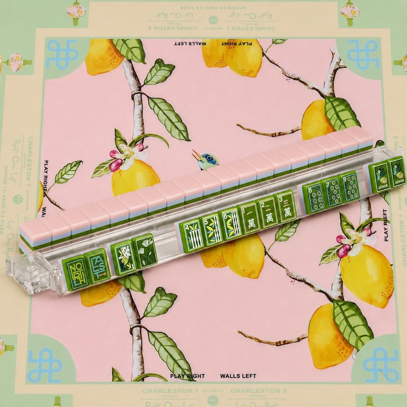 May include: A clear acrylic mahjong tile rack holding a row of tiles with pink backs and green fronts featuring various symbols, Chinese characters, and lemon illustrations, resting on a pink surface patterned with yellow lemons and green leaves.