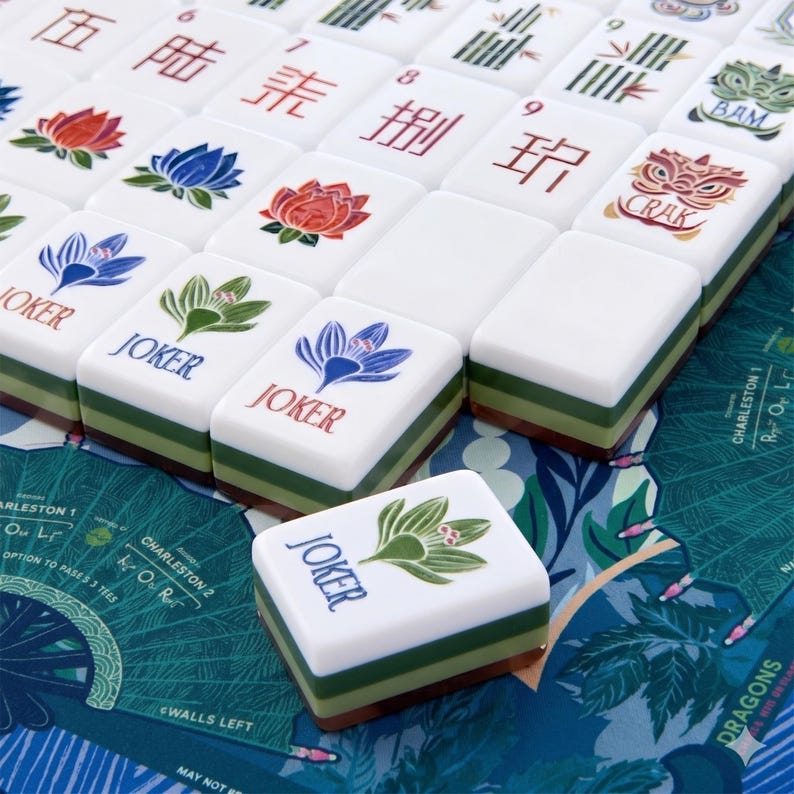 Puede incluir: Un conjunto de fichas de Mahjong blancas con bases superpuestas en verde y marr&oacute;n. Las fichas presentan ilustraciones coloridas de flores, caracteres chinos y la palabra JOKER en azul o rojo. Algunas fichas muestran el texto BAM o CRAK.