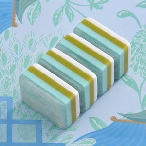 Puede incluir: Cuatro baldosas rectangulares dispuestas en fila, cada una con un dise&ntilde;o en capas que incluye una secci&oacute;n de color azul claro jaspeado, una franja de color verde oliva s&oacute;lido y una franja blanca s&oacute;lida. Tienen un acabado liso y brillante.