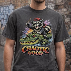 May include: Dark gray t-shirt featuring a raccoon wearing a red cap, riding a green alligator. The raccoon holds a stick, and the text "CHAOTIC GOOD" is displayed in orange and yellow. The design has a vintage, distressed look.
