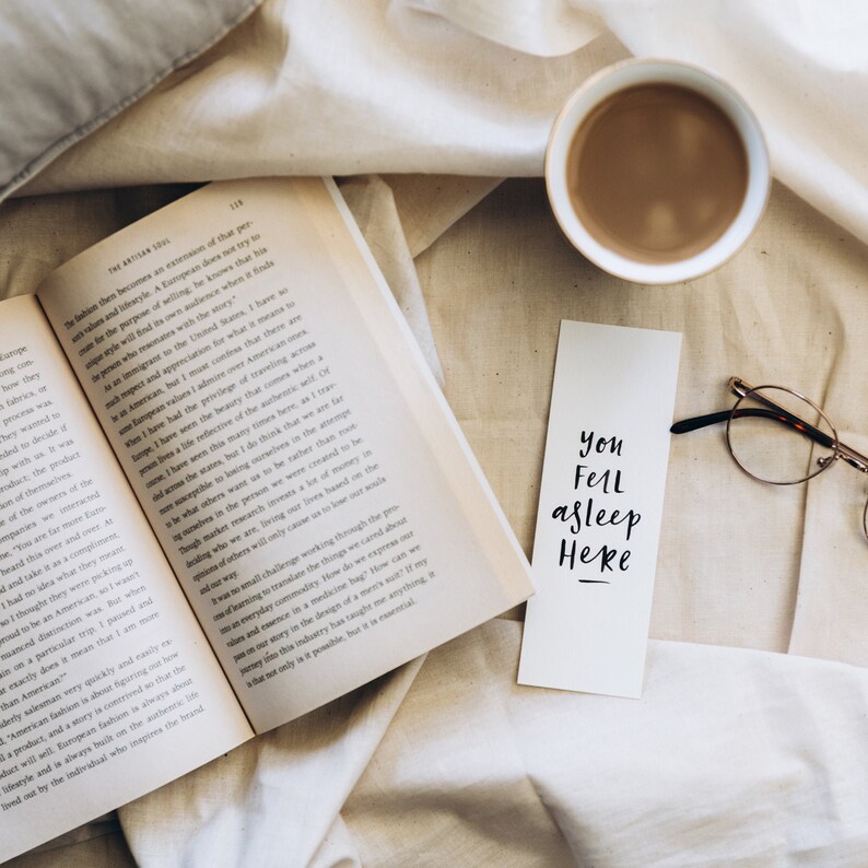 May include: A book, coffee cup, bookmark with "You Fell asleep Here", and spectacles on a white cloth. The image suggests a peaceful moment of reading and relaxation, with a warm beverage.