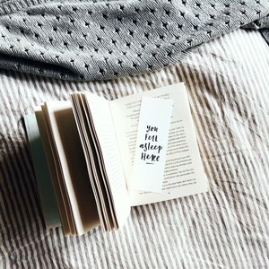 May include: An open book with a white bookmark featuring the text "You Fell Asleep Here" in black script. A grey sweater with a diamond pattern rests in the background, on a striped surface.