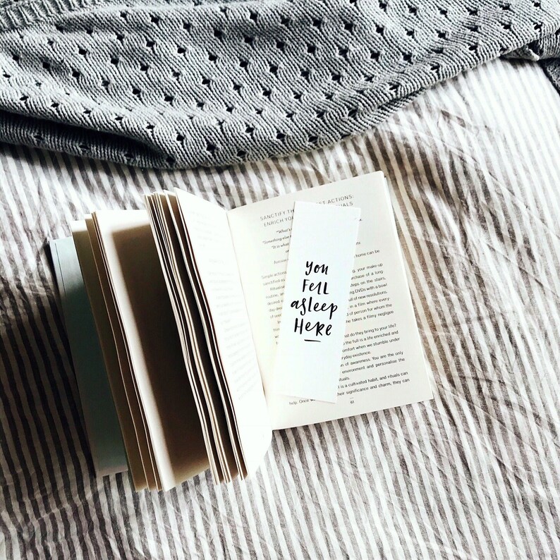 May include: An open book with a white bookmark featuring the text "You Fell Asleep Here" in black script. A grey sweater with a diamond pattern rests in the background, on a striped surface.