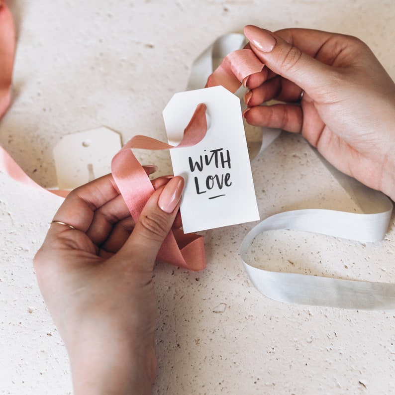 May include: A white gift tag with the words "WITH LOVE" in black script is held with pink ribbon. A second tag and white ribbon are also visible. The background is a light, textured surface.