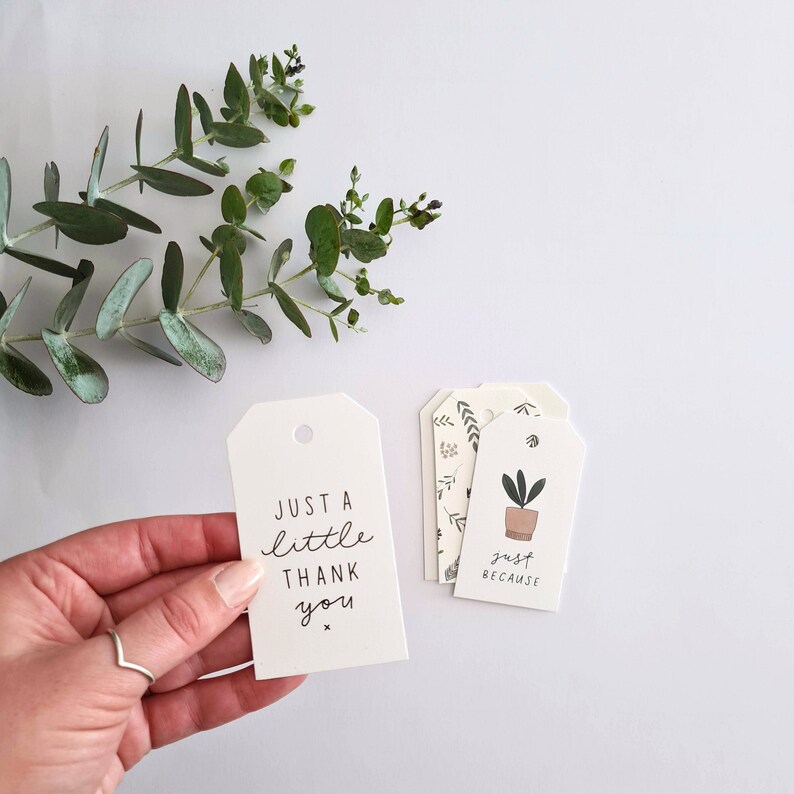 May include: White gift tags with black text and botanical illustrations. One tag reads "Just a little thank you." Other tags feature a potted plant and the words "just because." Green eucalyptus leaves are in the background.