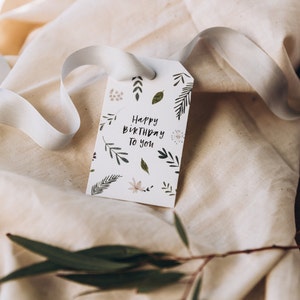 May include: A white gift tag with the text "Happy Birthday To You" and botanical illustrations. The tag is tied with a white ribbon and rests on a beige fabric surface. Green leaves and sprigs are scattered around the tag.
