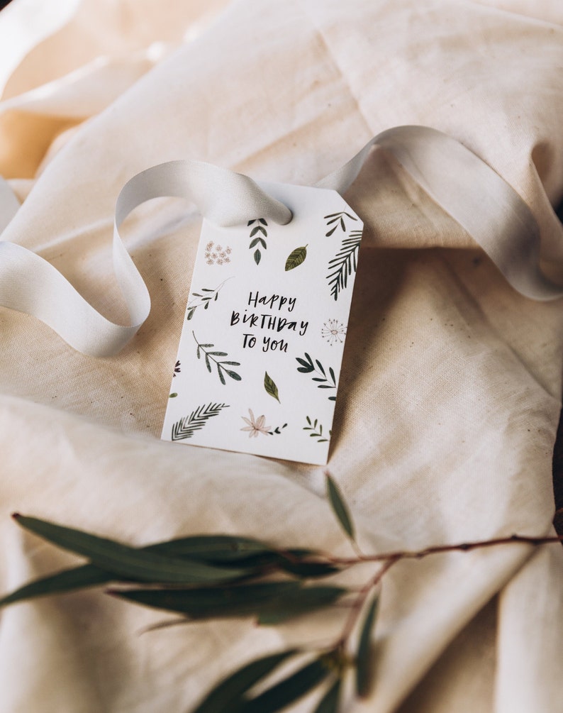 May include: A white gift tag with the text "Happy Birthday To You" and botanical illustrations. The tag is tied with a white ribbon and rests on a beige fabric surface. Green leaves and sprigs are scattered around the tag.